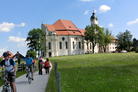 Cyclists and pedestrians travel past a historic chalet and church in Steingaden, Germany, on a sunny day.