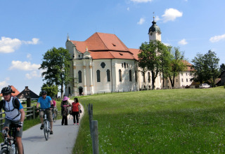 Cyclists and pedestrians travel past a historic chalet and church in Steingaden, Germany, on a sunny day.