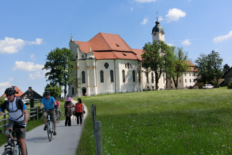 Cyclists and pedestrians travel past a historic chalet and church in Steingaden, Germany, on a sunny day.