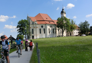Cyclists and pedestrians travel past a historic chalet and church in Steingaden, Germany, on a sunny day.