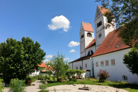 Garden and church with red roofs at Steingaden chalet, blue sky and trees, summer day in Germany.