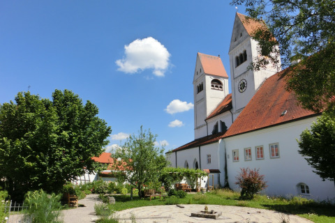Garden and church with red roofs at Steingaden chalet, blue sky and trees, summer day in Germany.