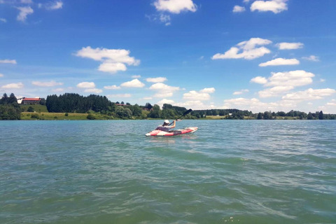 Person kayaking on a lake near Steingaden chalet in Germany, scenic countryside and clouds in view.