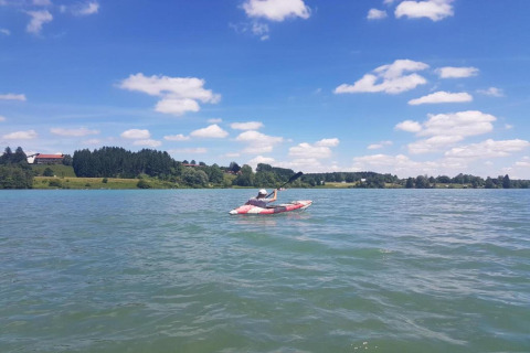 Person kayaking on a lake near Steingaden chalet in Germany, scenic countryside and clouds in view.