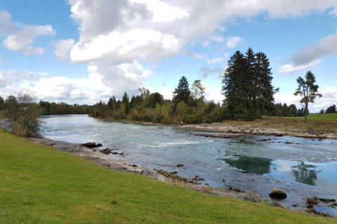 River landscape at Steingaden chalet in Germany with clear water, grassy banks, and trees under a cloudy sky.