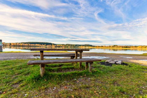 Picnic table and benches by a lake near Steingaden chalet in Germany, with peaceful nature and blue sky.