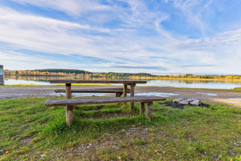 Picnic table and benches by a lake near Steingaden chalet in Germany, with peaceful nature and blue sky.