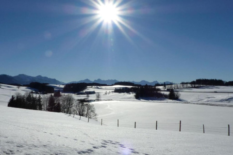 Sunny winter day with snow-covered fields and distant mountains at chalet Steingaden in Germany, blue sky.