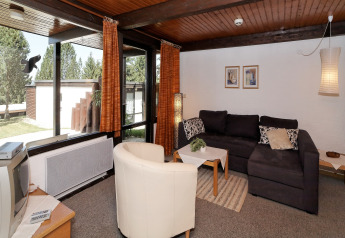 Living area at Holiday park «Jägerwiesen 1» in Germany with sofa, armchair, TV, and large windows.