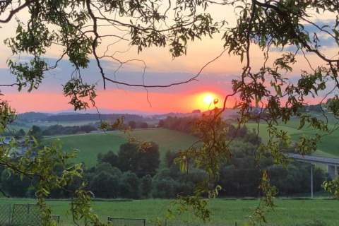 Sonnenuntergang über der hügeligen Landschaft bei Holiday park «Jägerwiesen 1», mit Bäumen im Vordergrund.