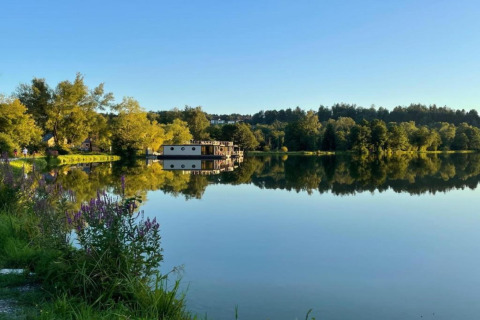Reflection of Holiday park «Jägerwiesen 1» lodge on a calm lake in Germany, surrounded by trees and flowers.