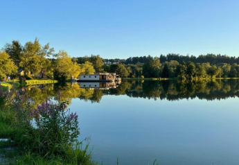 Reflection of Holiday park «Jägerwiesen 1» lodge on a calm lake in Germany, surrounded by trees and flowers.