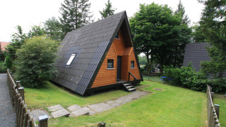 A-vormig houten chalet Type A1 - Nurdachhaus met zwarte dakpannen, veranda en omringd door groen gras.