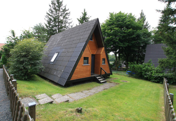 A-vormig houten huisje Type A1 - Nurdachhaus, met donker dak, veranda en een groene tuin met bomen.