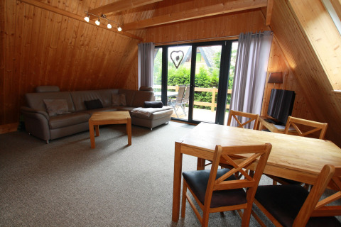 Living room in a wooden lodge with a grey couch, dining table, chairs, and balcony view to greenery.