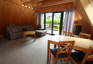 Woonkamer in een houten lodge met grijze bank, eettafel, stoelen en uitzicht op balkon met groen.