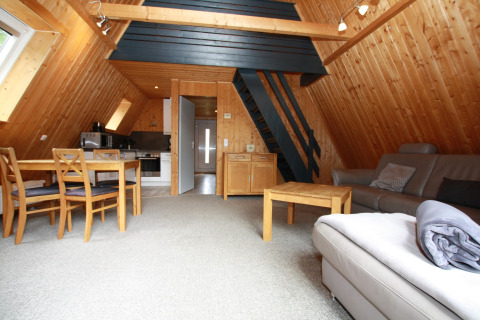 Sfeervol lodge-interieur met houten wanden, open keuken, eettafel, bank en trap naar de zolder.