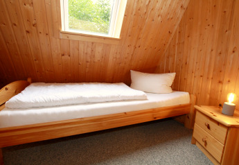 Petite chambre dans un lodge avec lit simple, murs en bois, fenêtre, table de nuit et lampe allumée.