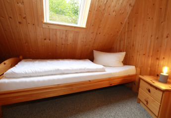 Petite chambre dans un lodge avec lit simple, murs en bois, fenêtre, table de nuit et lampe allumée.