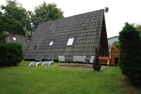 Foto van een A-frame lodge met ligstoelen, bankjes, vuurkorf en houten terras op een groen gazon.