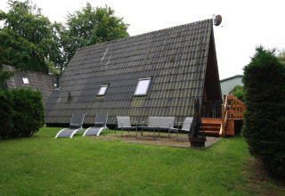 Photo of an A-frame lodge with sun loungers, chairs, fire pit, and wooden terrace in a grassy yard.