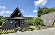 A-frame wooden lodge house with green windows and garden, photographed in Germany on a sunny day.