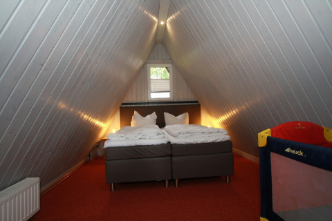Slaapkamer in een A-vormige lodge in Duitsland met twee bedden, rood tapijt en kinderbedje in hoek.