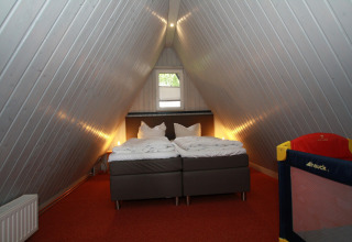 Slaapkamer in een A-vormige lodge in Duitsland met twee bedden, rood tapijt en kinderbedje in hoek.