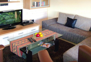 Woonkamer in vakantiepark Öfingen, Duitsland, met sofa, glazen tafel, fruit en tv in een chalet.