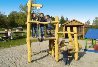 Children playing on a wooden playground at Holiday village Öfingen, with adults and cabins in the background.