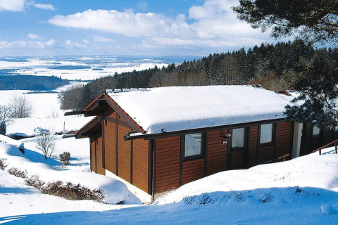 Chalet cubierto de nieve en Holiday Village Öfingen, Alemania, rodeado de colinas y paisaje invernal.