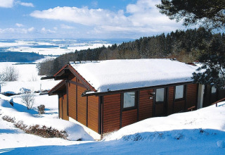 Chalet cubierto de nieve en Holiday Village Öfingen, Alemania, rodeado de colinas y paisaje invernal.