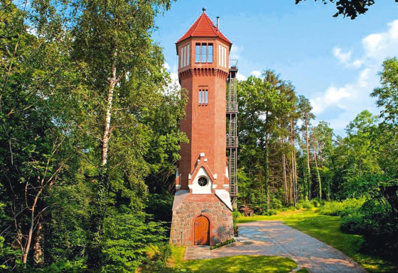 Udsigt til den charmerende Water tower 75 sqm-chalet, omgivet af frodige grønne træer under blå himmel.