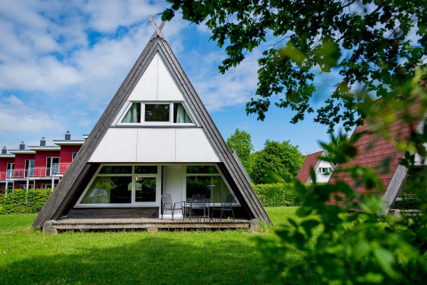 A photo of an A-frame lodge house with large windows and patio furniture on a green lawn with trees around.