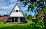 A photo of an A-frame lodge house with large windows and patio furniture on a green lawn with trees around.
