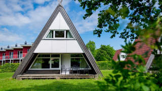A photo of an A-frame lodge house with large windows and patio furniture on a green lawn with trees around.