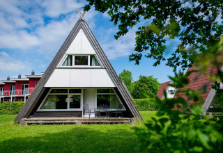 Foto van een A-vormig chalet met grote ramen en een terras op het gras, omgeven door bomen en natuur.