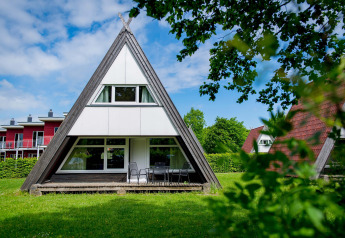 A photo of an A-frame lodge house with large windows and patio furniture on a green lawn with trees around.