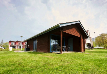 Et moderne træhus-lodge med terrasse og havemøbler på en grøn græsplæne med flere hytter i baggrunden.