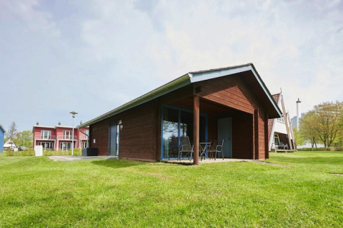 Moderno lodge in legno con patio coperto e sedie da esterno, circondato da prato verde e altre case in lontananza.