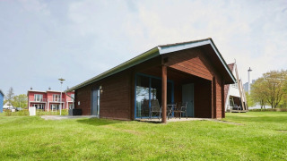 Et moderne træhus-lodge med terrasse og havemøbler på en grøn græsplæne med flere hytter i baggrunden.