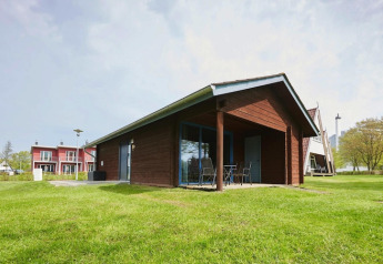 A modern wooden lodge with a covered patio and outdoor chairs, set on green grass with other lodges nearby.