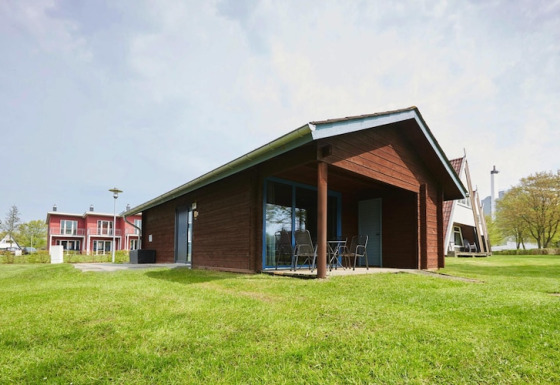 Moderno lodge in legno con patio coperto e sedie da esterno, circondato da prato verde e altre case in lontananza.