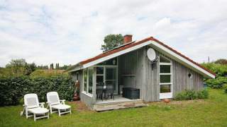 Holiday home in Hünning with wood exterior, sloped red roof, lawn, and two white lounge chairs outside.