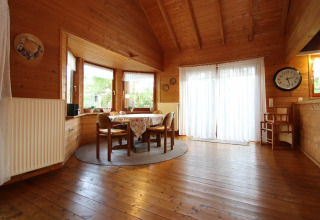 Cozy dining area in a wooden lodge with round table, chairs, bay windows, and white curtains.