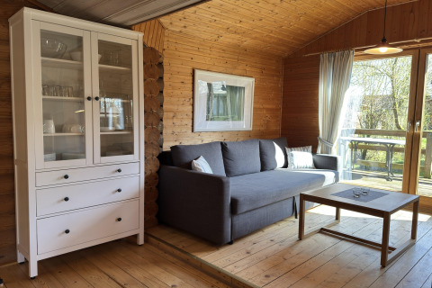 Woonkamer in een houten lodge in Duitsland met een grijze sofa, witte kast en zicht op zonnig terras.