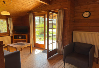 Cozy lodge interior with open door to garden, armchairs, sofa, TV, and wood decor, Type C House in Germany.