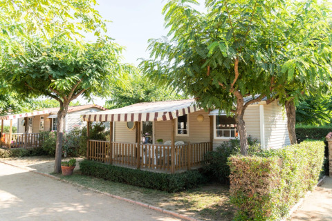 Casa móvil en Camping del Mar con porche y toldo a rayas, rodeada de árboles y arbustos verdes.
