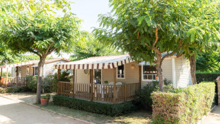 Camping del Mar stacaravan met gestreept zonnescherm, omringd door bomen en struiken in de zomer.