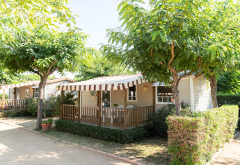 Casa móvil en Camping del Mar con porche y toldo a rayas, rodeada de árboles y arbustos verdes.